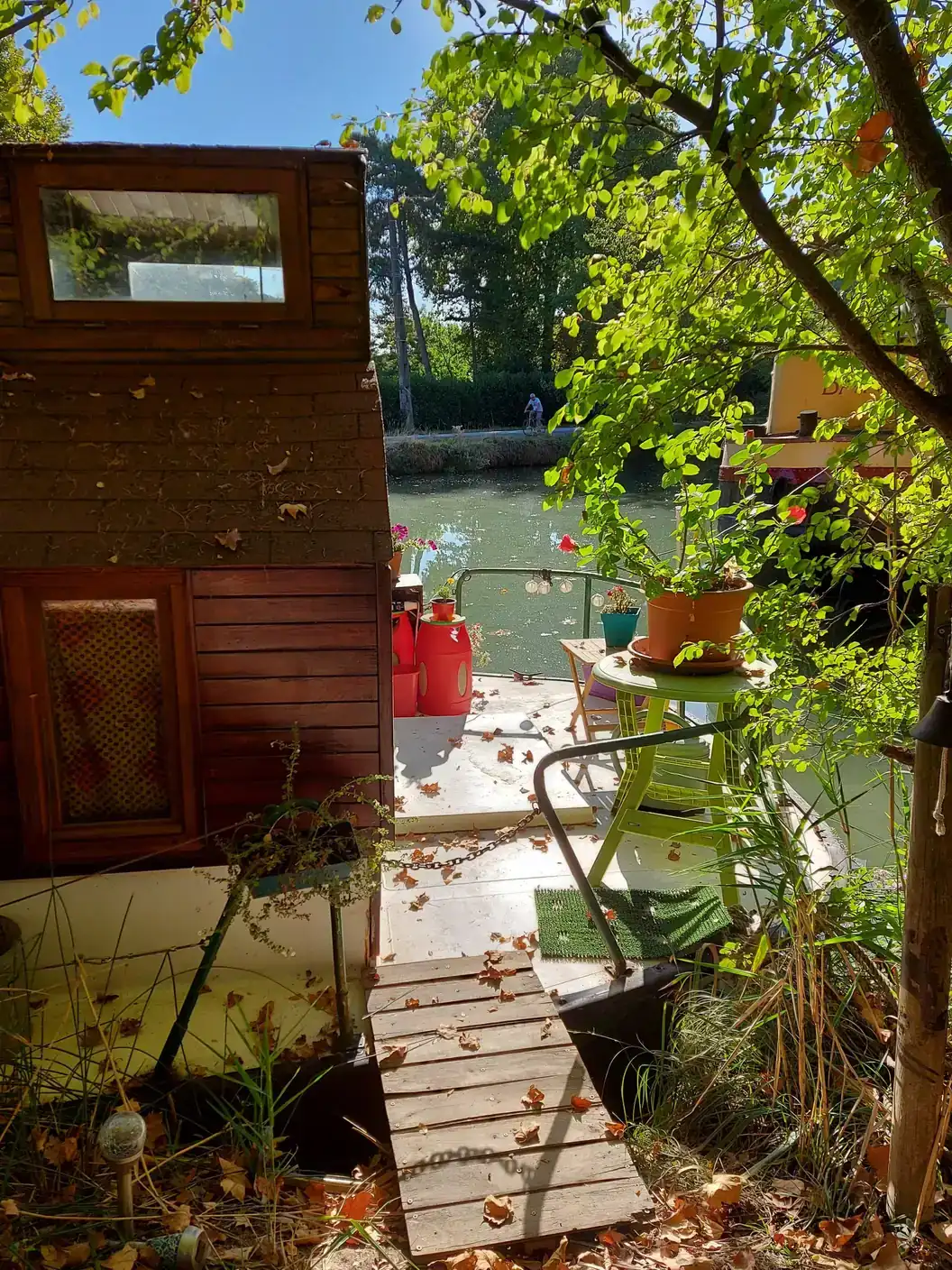 Sur le Canal du Midi à Ramonville-Saint-Agne, une péniche en bois et son ponton fleuri, avec un cycliste sur la berge d'en face.
