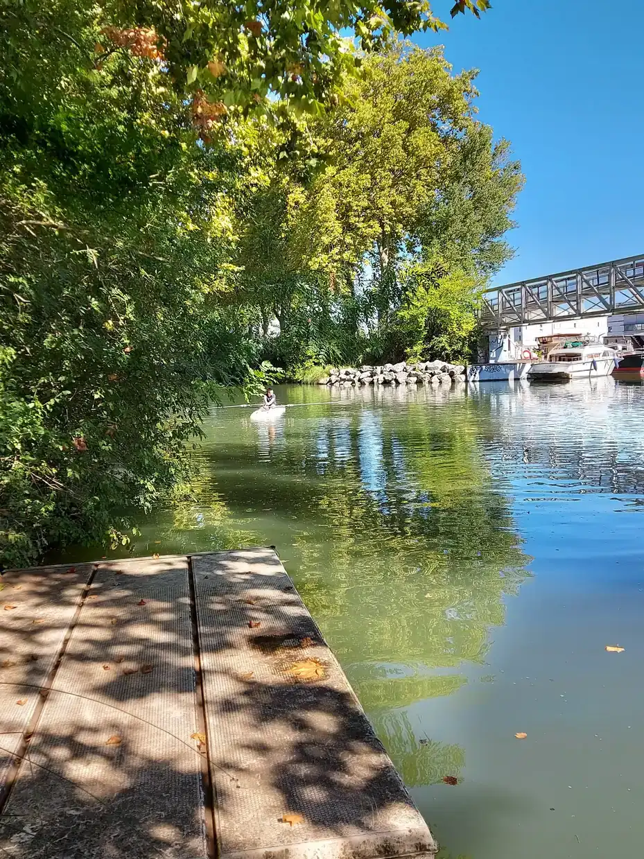 Un ponton d'aviron ombragé sur le Canal du Midi à Ramonville-Saint-Agne, avec une passerelle et des bateaux amarrés au fond.
