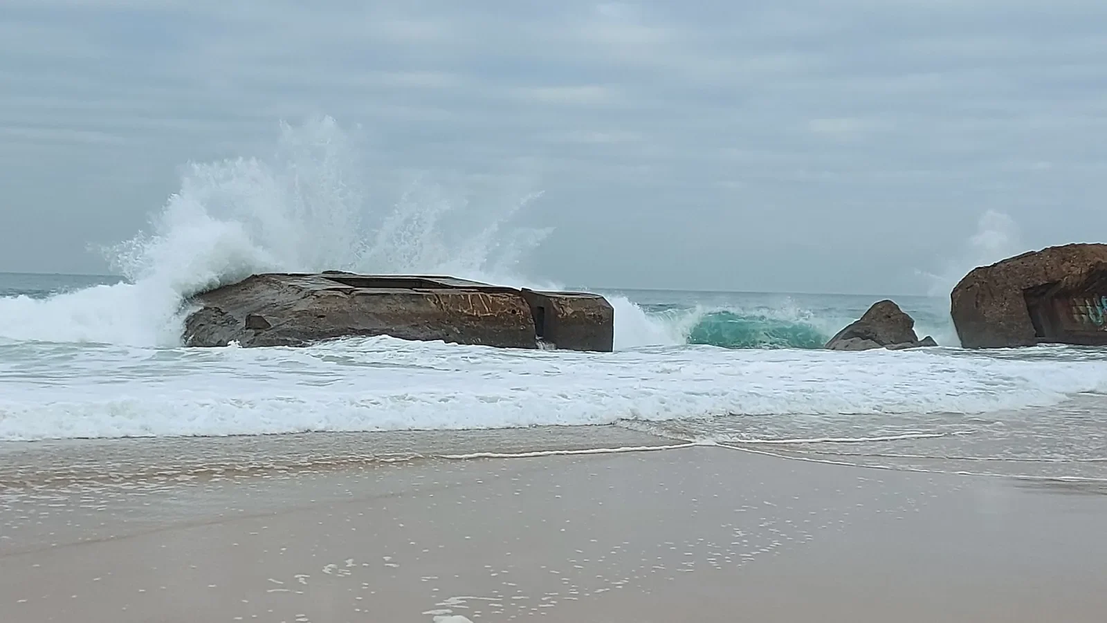 Une vague s'écrase sur un blockhaus échoué à Capbreton, dans un jaillissement d'embruns, sous un ciel gris.
