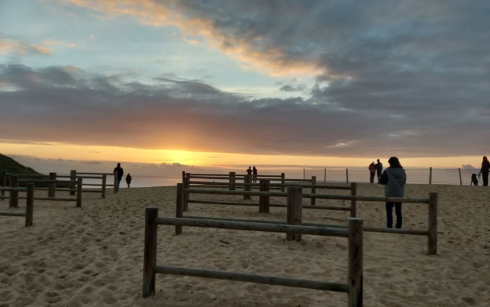 Au coucher du soleil, des promeneurs s'attardent entre des barrières en bois sur la dune de Capbreton, face à l'océan.