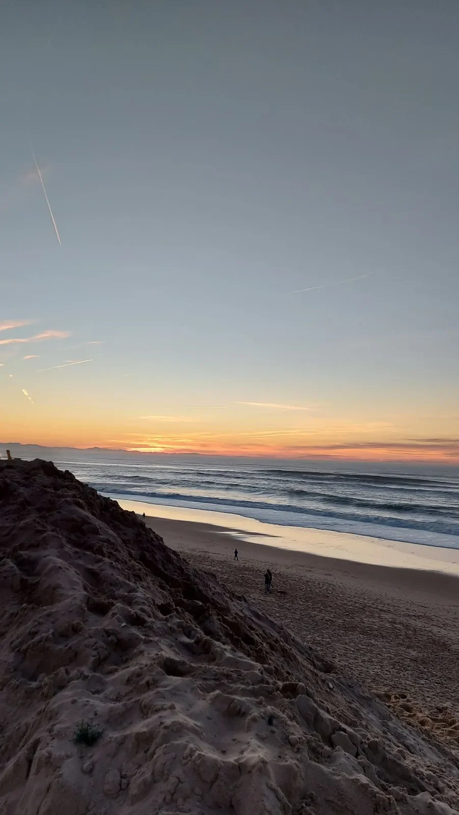 Le soleil couchant sur l'océan à Capbreton, vu depuis une dune de sable, avec quelques promeneurs en silhouette sur la plage.