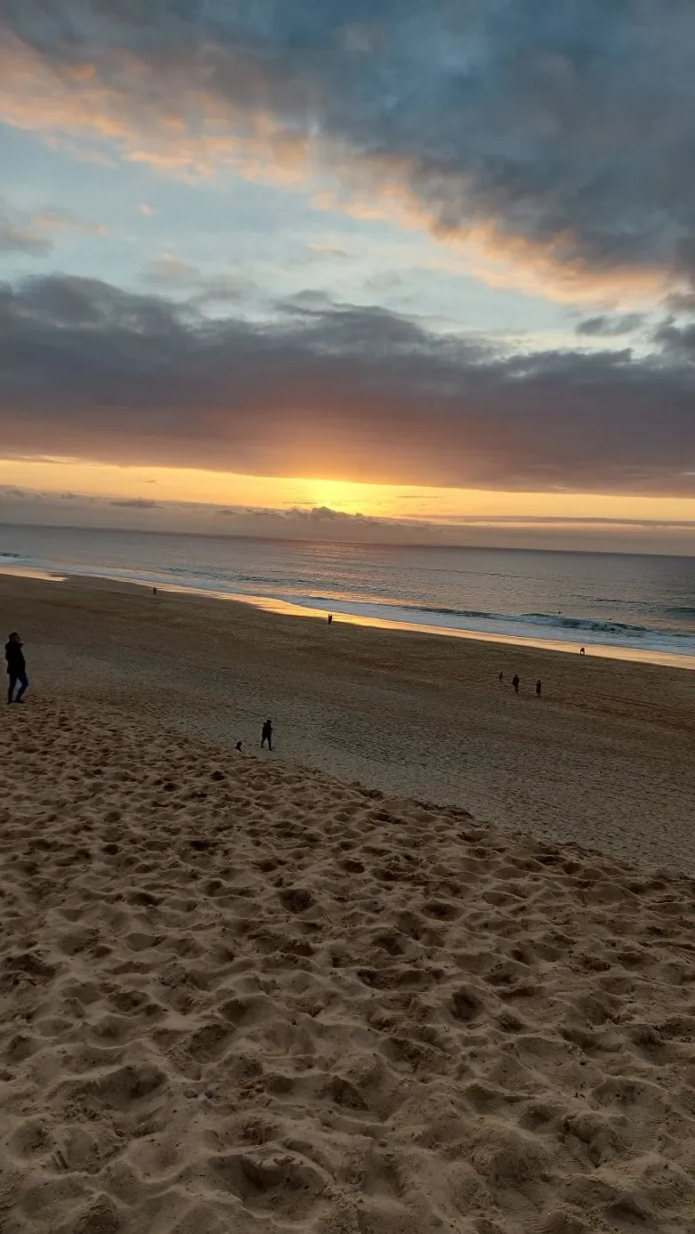 Le soleil descend sous un ciel de nuages roses et dorés, vu depuis la dune de Capbreton, avec quelques promeneurs sur la plage.