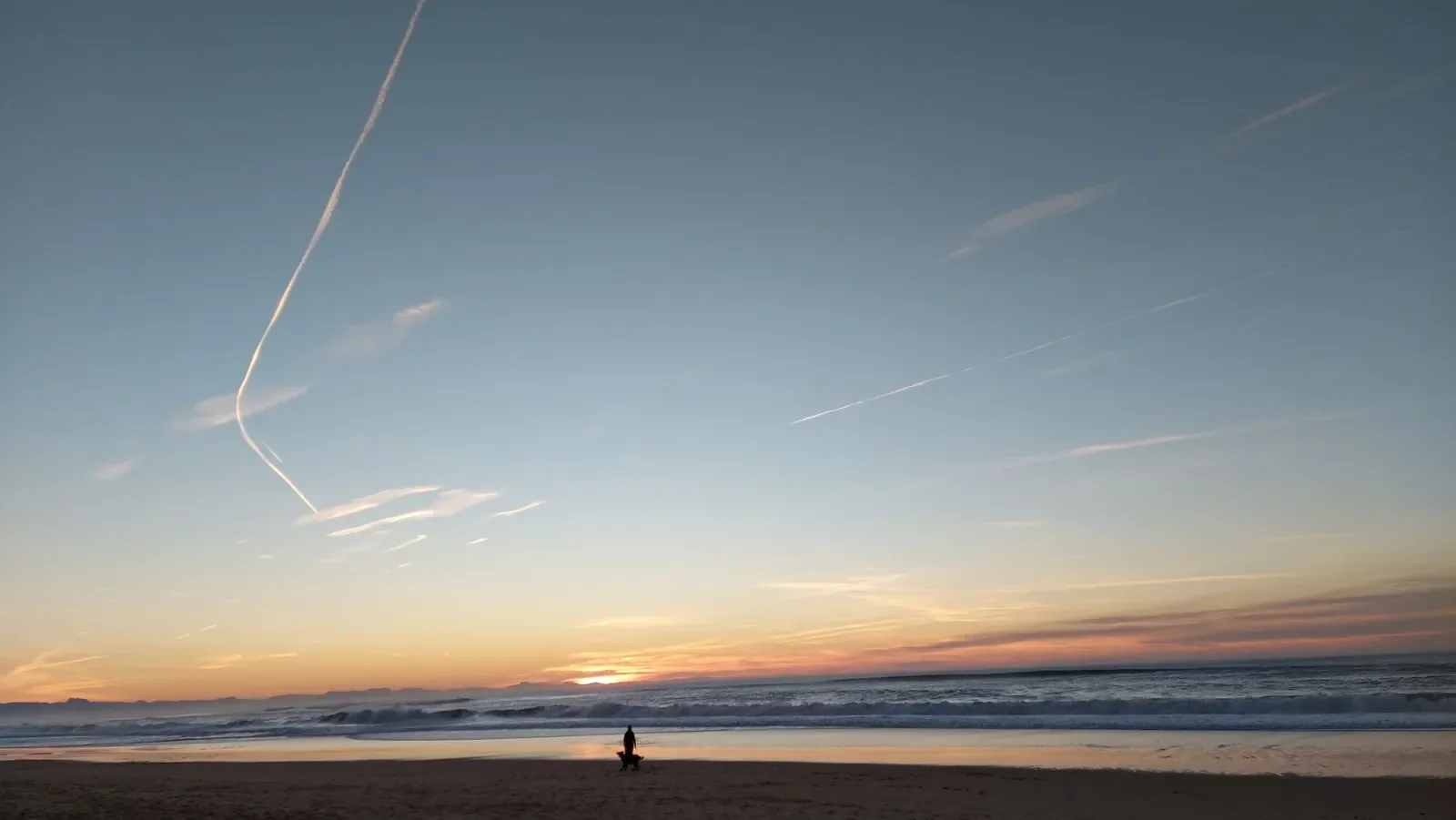 Une silhouette et son chien face au soleil couchant à Capbreton, sous un ciel strié de traînées d'avion.