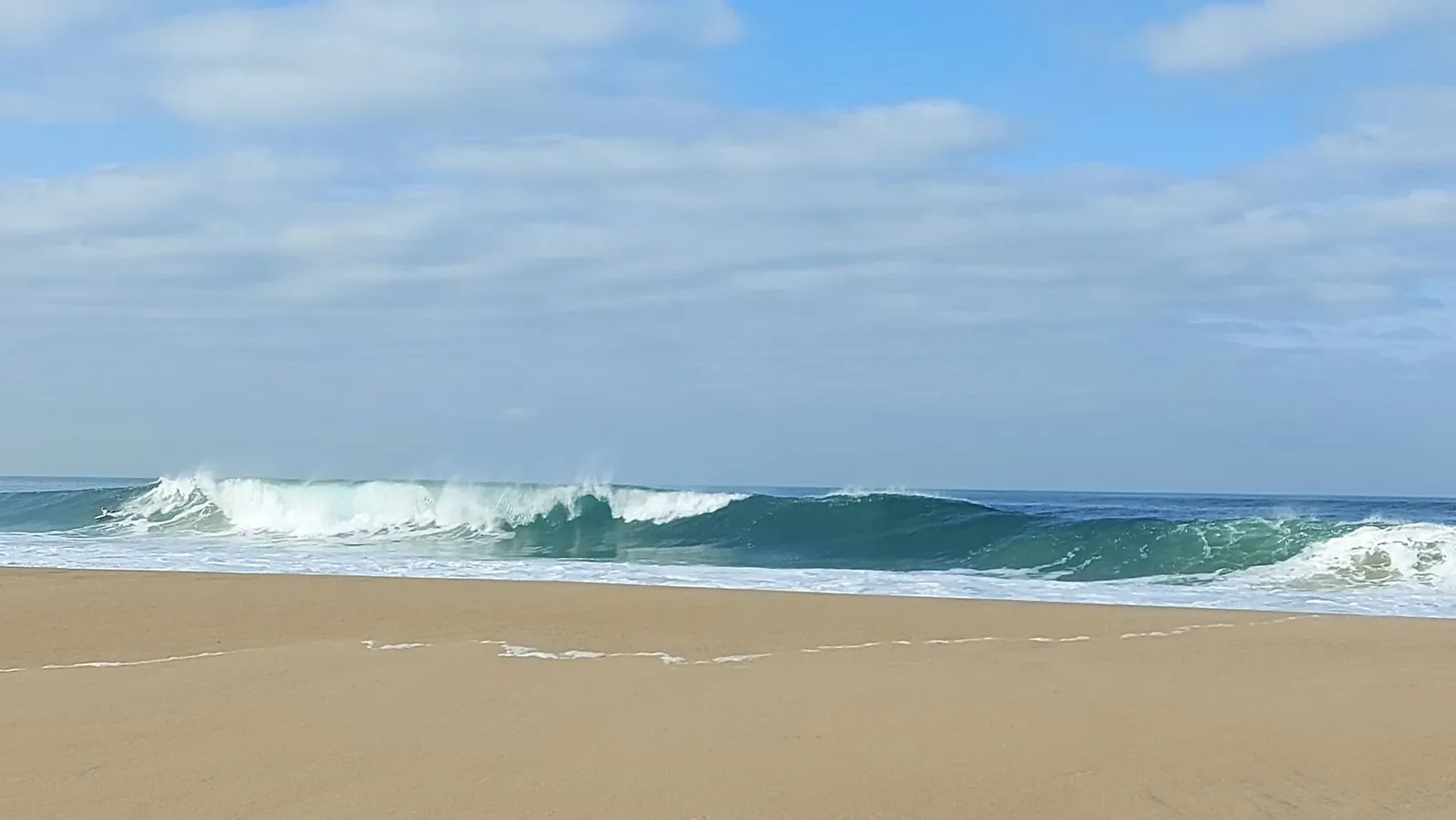 Une vague turquoise déferle sur la plage de Capbreton, sous un ciel bleu parsemé de nuages, un matin de novembre.