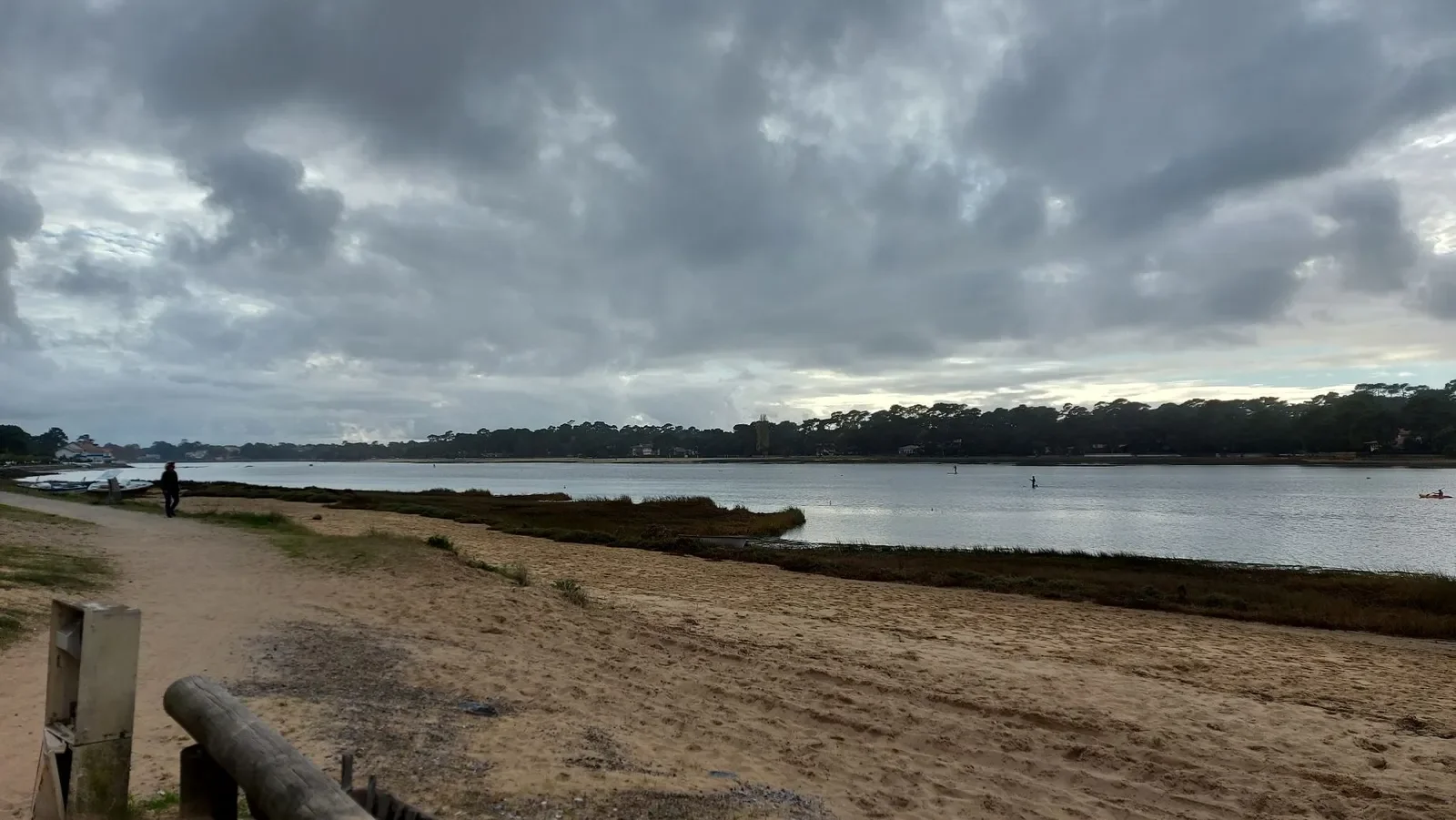 Des pratiquants de paddle sur le lac marin d'Hossegor, sous un ciel de nuages, avec un sentier au bord de l'eau.