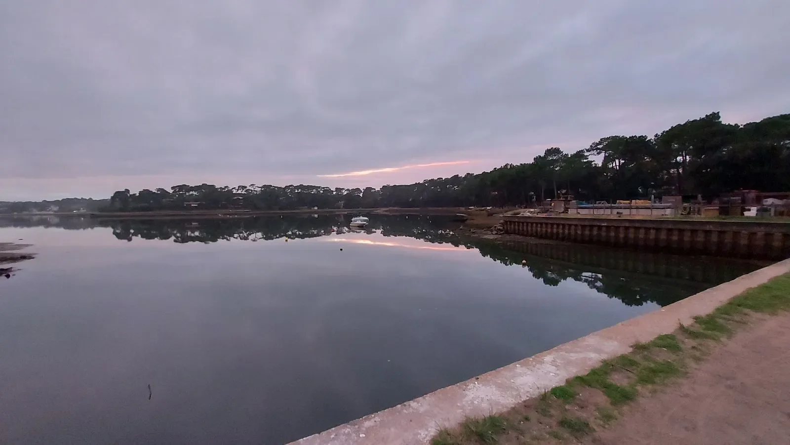 Le lac marin d'Hossegor, calme au crépuscule, reflète les pins et un trait de lumière rose, le long d'un quai.