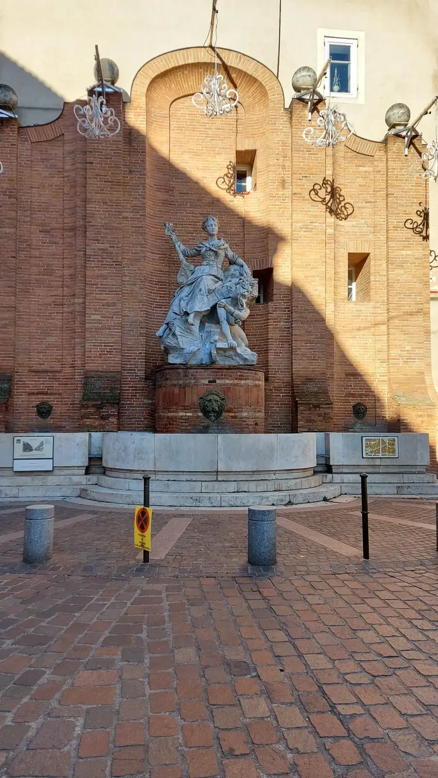 Fontaine avec une statue de femme et un lion devant un mur de briques roses, dans une rue pavée de Toulouse