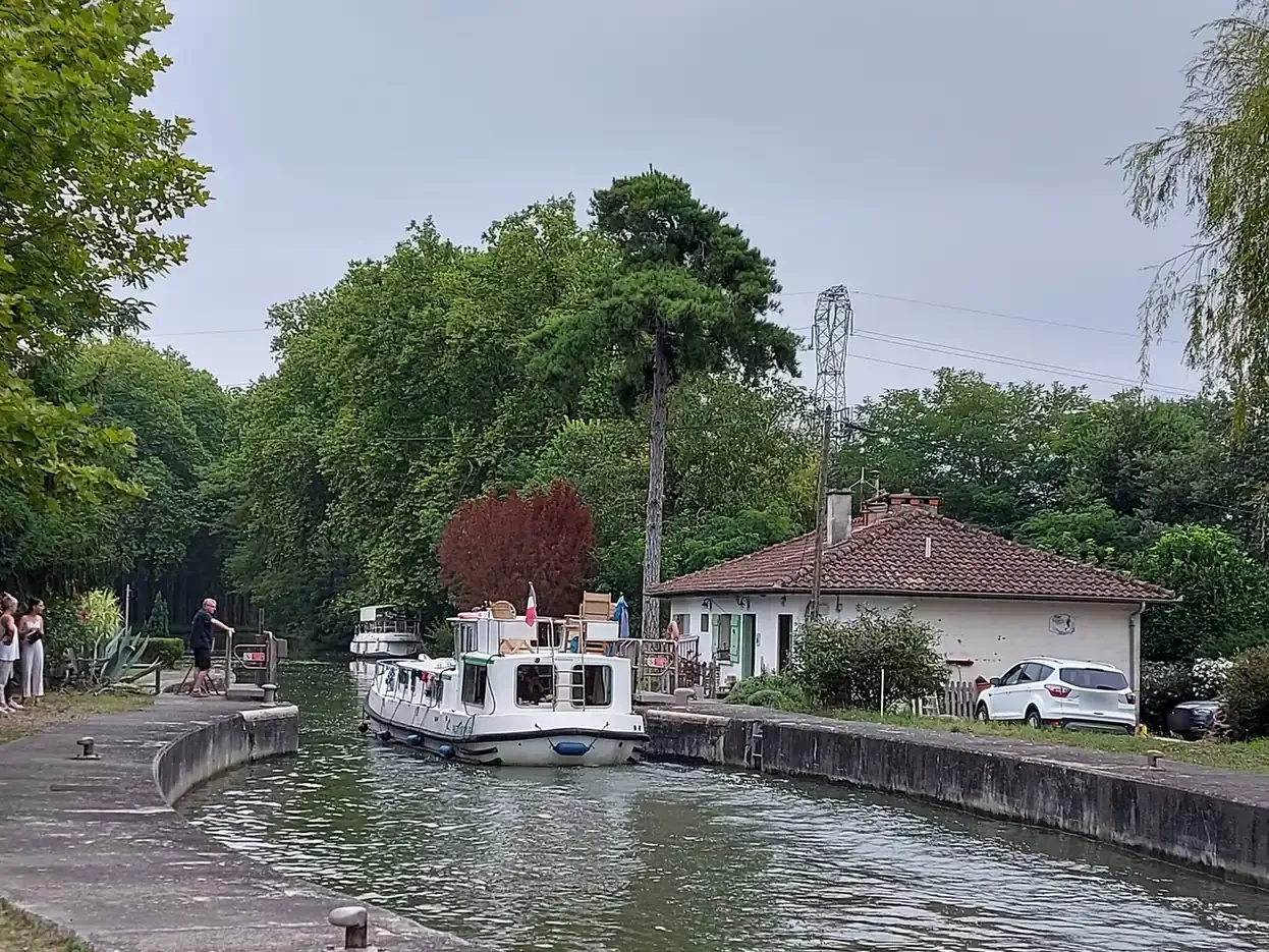Péniche blanche sortant d'une écluse du canal du Midi, avec la maison éclusière et des promeneurs qui regardent.