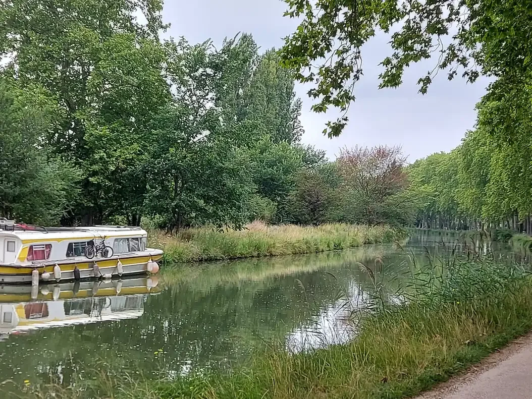 Péniche blanche et jaune amarrée, reflétée dans l'eau calme du canal du Midi, avec un vélo posé sur le toit