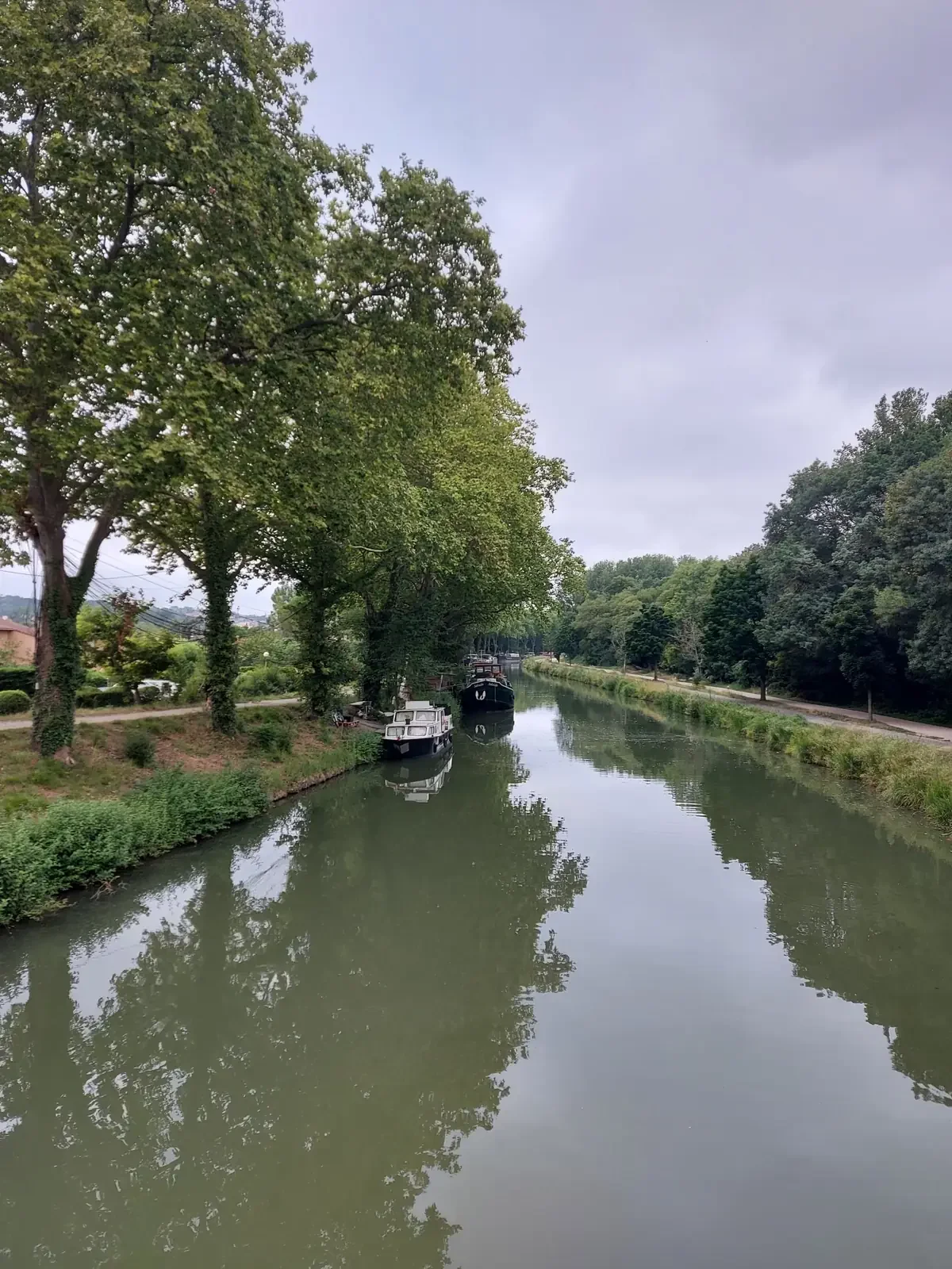 Le canal du Midi sous un ciel gris, avec des péniches amarrées et le reflet des arbres dans l'eau