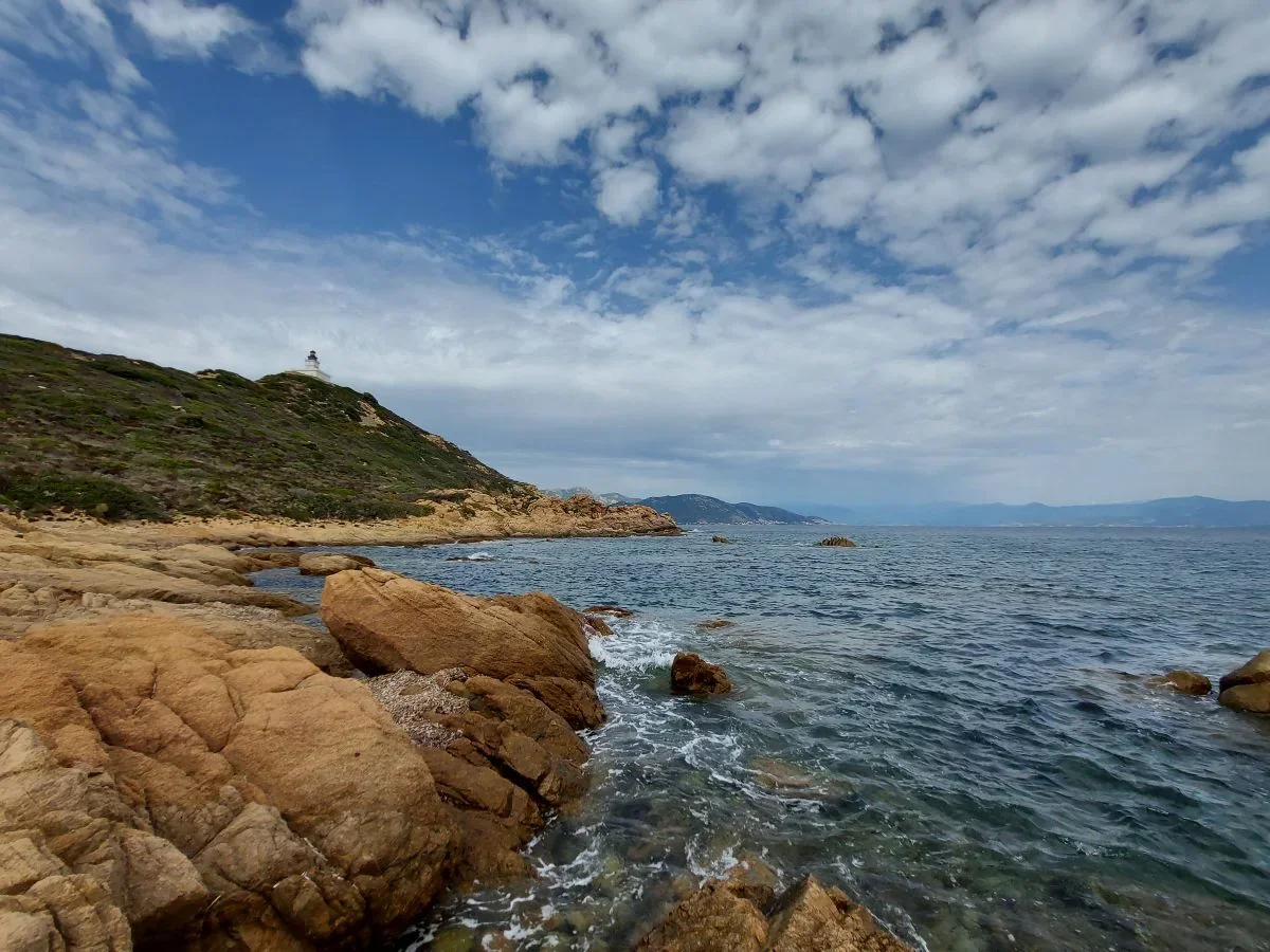 Rochers ocre dans une crique, mer transparente et le phare au loin